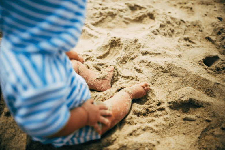 Baby in blue striped outfit sitting on sandy beach and playing with the sand
